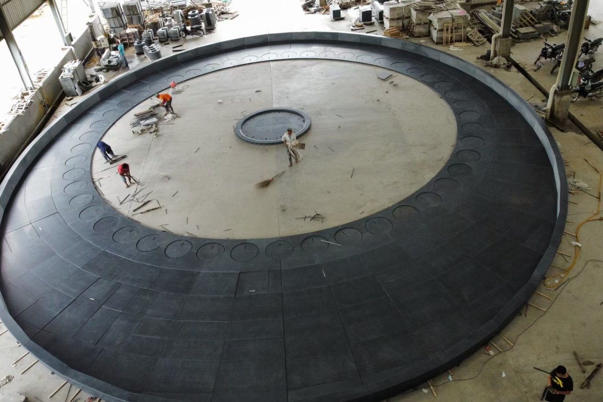 A very large circular dark stone floor assembly being dry-laid full size on the factory floor, with workers for scale